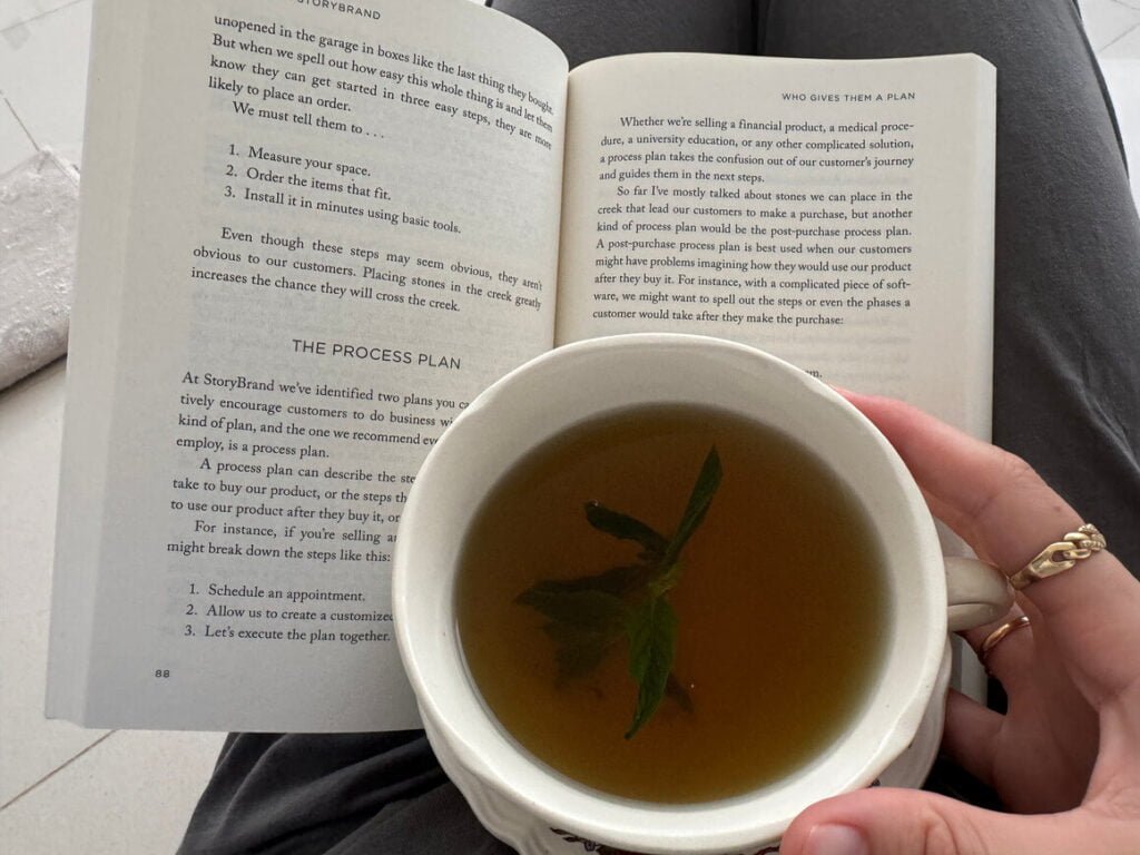 A girl sits holding a cup of sinus tea with mint with an open book on her lap.