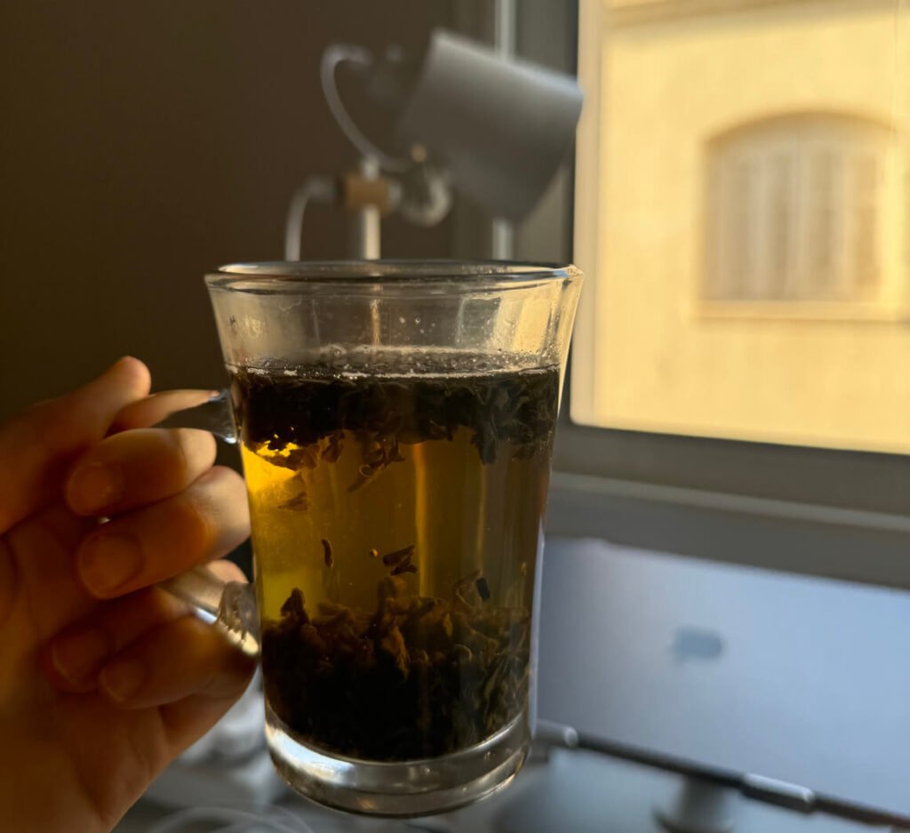 girl holding a cup of tea with infused leaves with a window and a macbook in the background